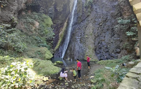 Pemandangan air terjun Curug Song yang dikelilingi hutan pinus hijau dengan latar belakang pegunungan dan suasana asri yang menyejukkan.